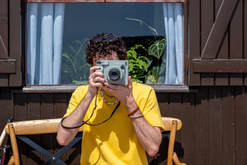 A person in a yellow shirt is seated in front of a window with white curtains and plants. They hold a grey instant camera up to their face, blocking their features. The scene is sunny, and the person wears sunglasses on their head, captured against a wooden structure.