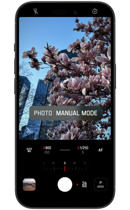 A smartphone screen displaying a photo of blooming pink flowers with a building in the background. The screen is in camera manual mode showing various settings such as ISO, shutter speed, white balance, and focus.