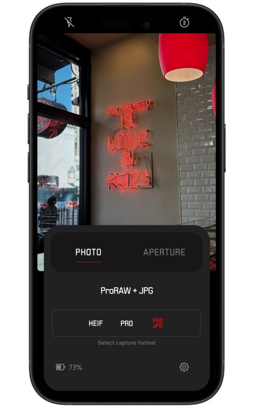 A smartphone screen displays a photograph taken in a cafe. The image shows a neon sign on a white tiled wall that reads, "All you need is love & pizza." The phone interface indicates options for capture format selection, with "ProRAW + JPG" currently chosen.