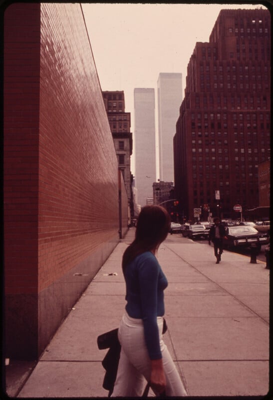 A person with long hair wearing a blue top and light-colored pants walks along a sidewalk in an urban area. The street is bordered by a tall brick wall on one side and several high-rise buildings on the other. The Twin Towers are visible in the background.