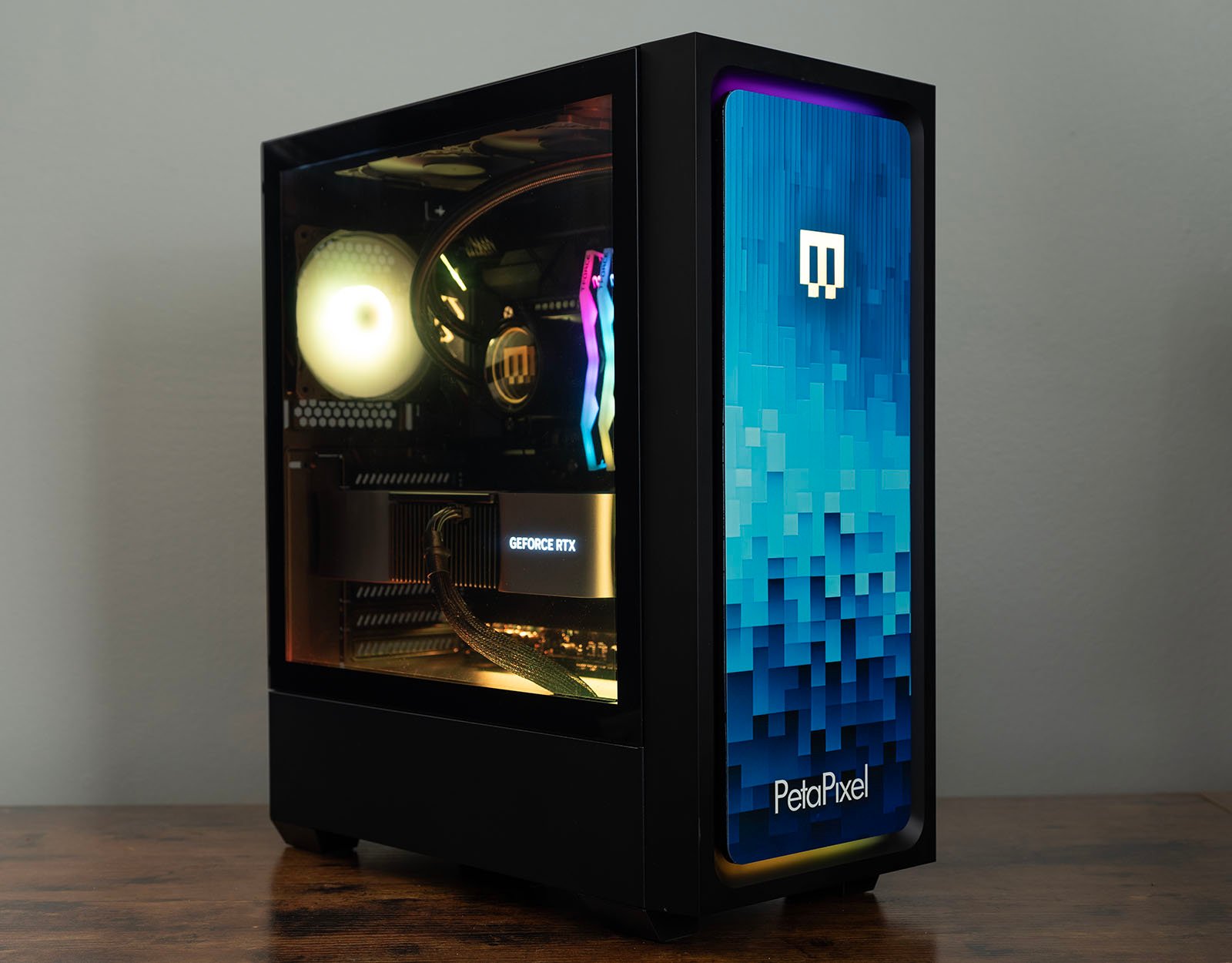 A black desktop computer tower with a transparent side panel revealing internal components, including a GeForce RTX graphics card. The front panel has a blue pixelated design with a white logo and "PetaPixel" text, illuminated with RGB lighting.
