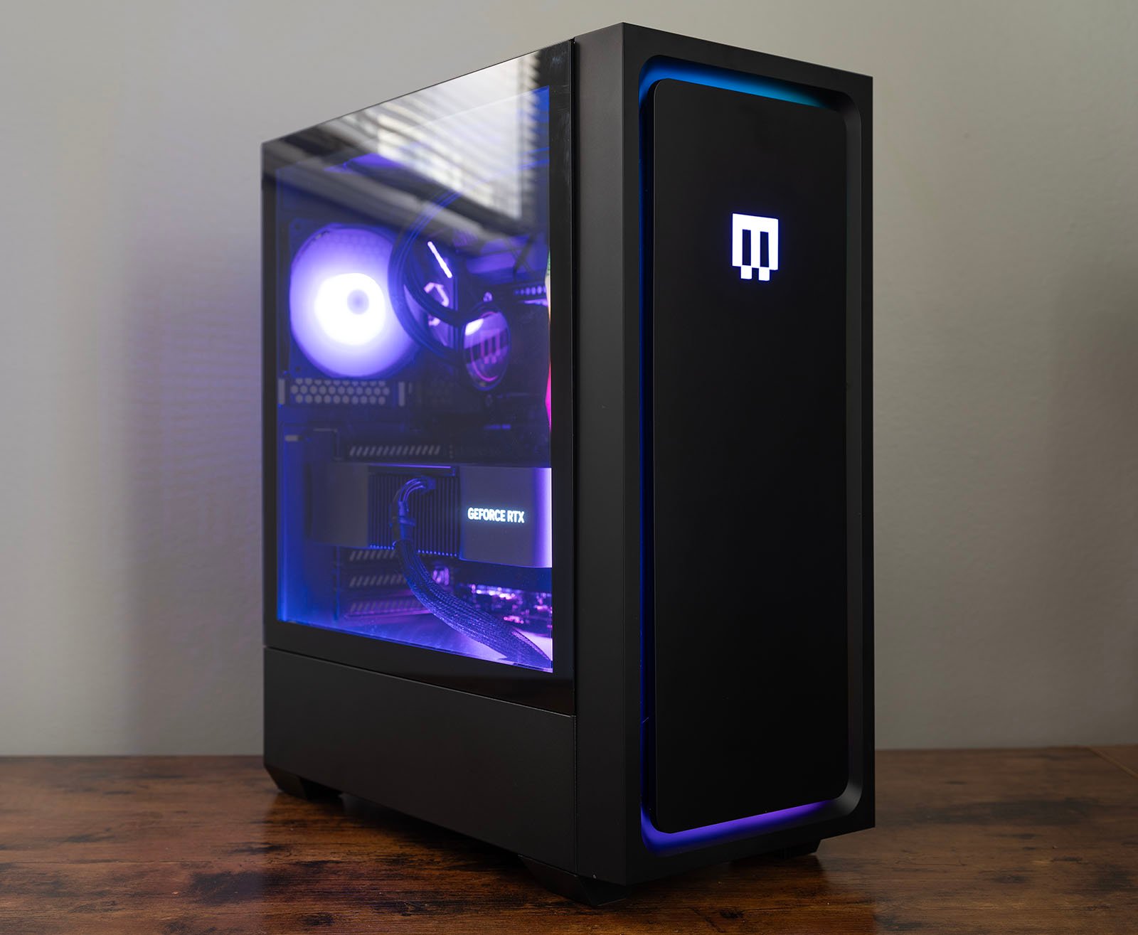 A sleek, black gaming PC tower with a side glass panel is shown on a wooden surface. The interior is illuminated with blue and purple LED lights, showcasing the components, including a GeForce RTX graphics card. The front panel features a glowing logo.