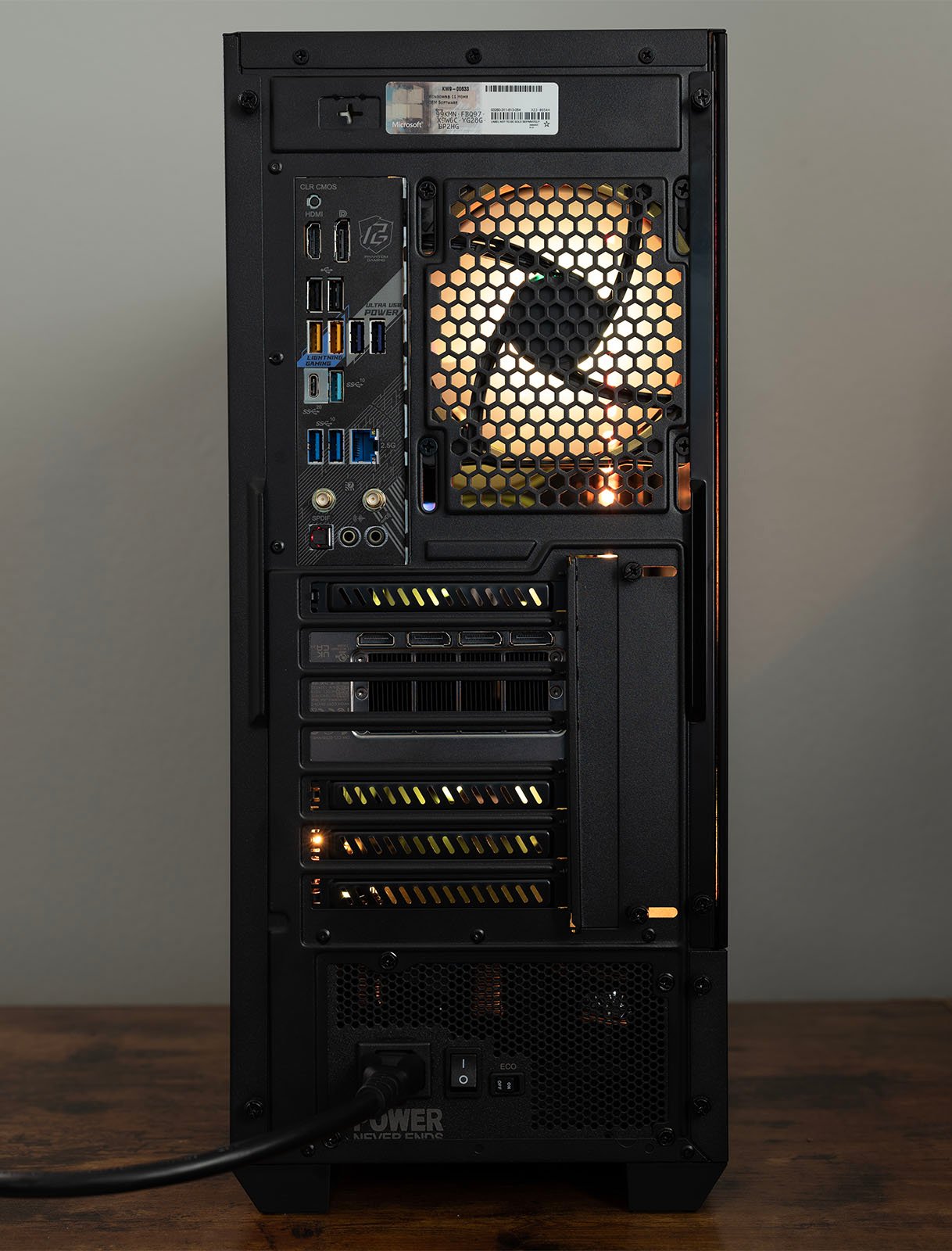 The image shows the rear view of a desktop computer tower featuring various ports including USB, Ethernet, HDMI, and audio jacks. A cooling fan is visible through the mesh panel, and the power supply is connected to the power cord at the bottom.