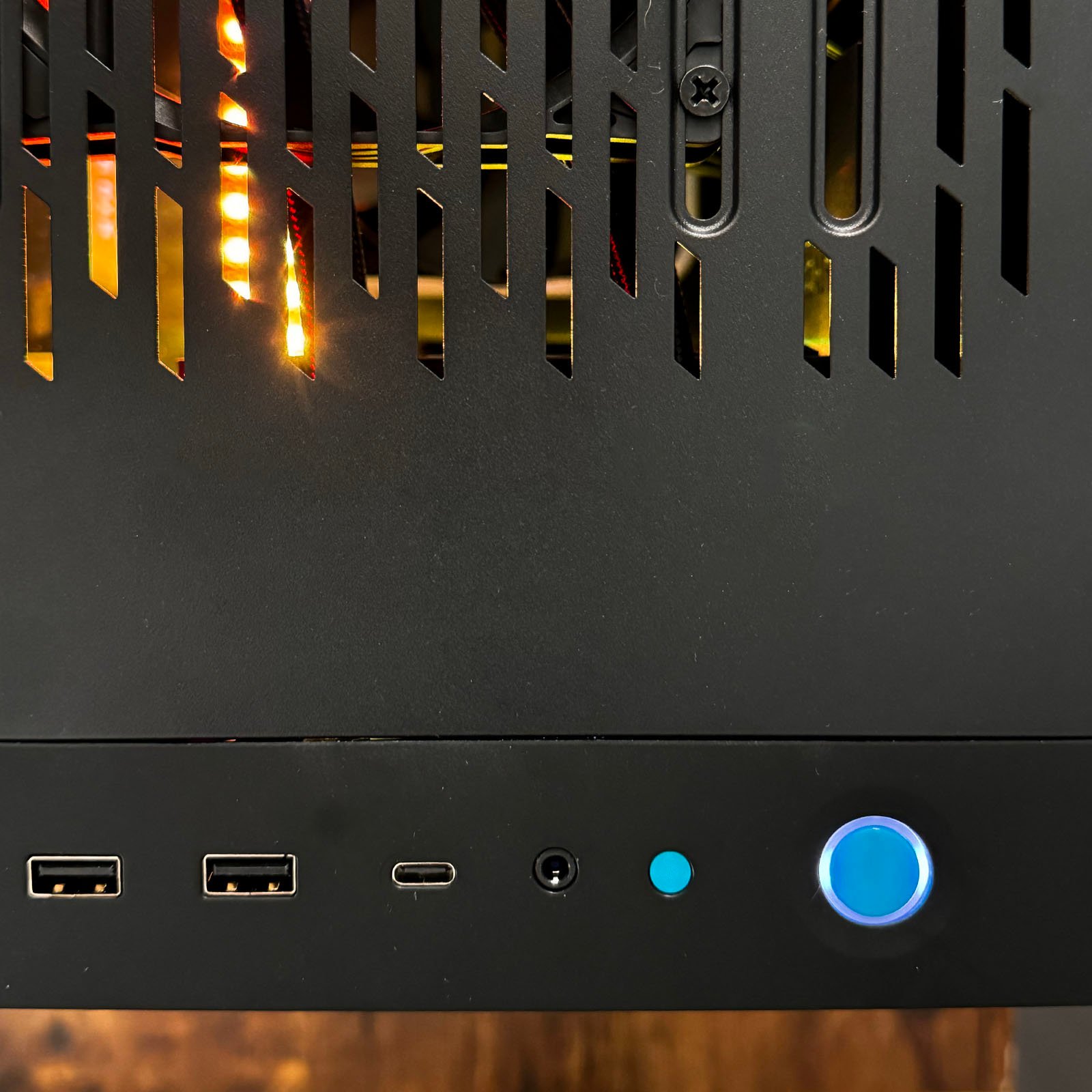 Close-up of a computer casing highlighting ventilation slits on the top half and multiple ports at the bottom. Ports include two USB-A ports, one USB-C port, a headphone jack, a small blue button, and a larger illuminated blue power button.
