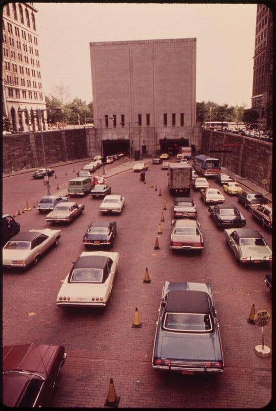 A vintage photograph shows numerous cars lined up in multiple lanes, waiting to enter a large tunnel surrounded by tall buildings in an urban setting. Traffic cones guide the vehicles. Trees and other buildings are visible in the background.