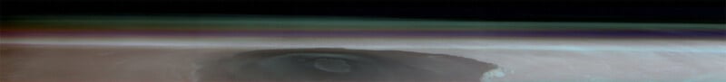 An ethereal image showing a multicolored atmospheric layer above the Martian surface. The thin, wispy clouds and haze create a gradient of colors, transitioning from green to blue. Below, a large crater with a central depression is visible on the planet's surface.