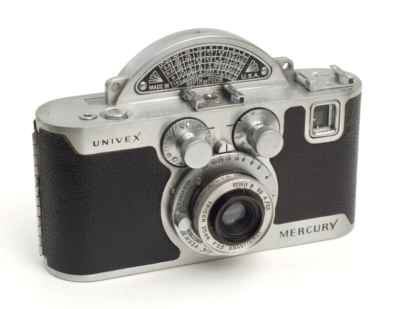 A vintage Univex Mercury II camera with a distinctive half-moon shaped exposure dial on top. The camera features a silver metal body with black textured grips and a prominent lens in the center. Various knobs and dials are present for adjustments.
