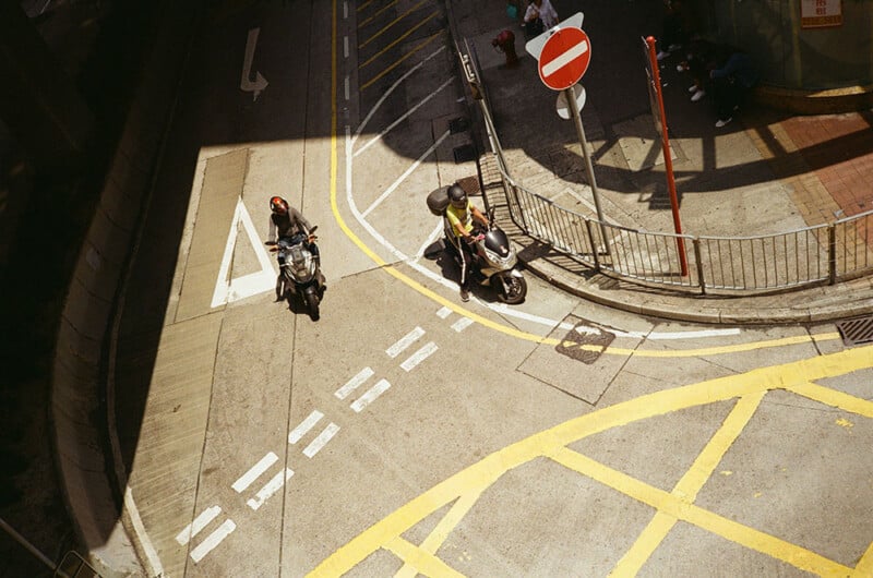 Aerial view of two motorcyclists stopped at a T-intersection on a city street. One rider is on the left side, and the other is by a "no entry" sign on the right side. The road markings include pedestrian crossing lines and yellow grid lines for traffic control.