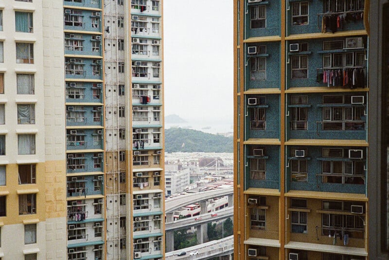 View of a cityscape through a narrow gap between two high-rise residential buildings, one in tan and white and the other in yellow and teal. Visible in the background are other buildings, hills, and the sea under a cloudy sky.