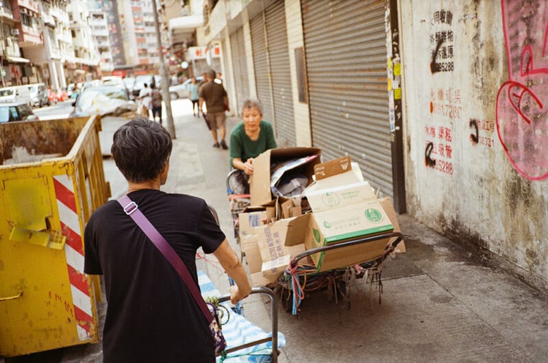 Two elderly people push carts loaded with cardboard boxes down a narrow street. The person in the foreground wears a black shirt and pushes a cart with a purple shoulder strap bag. The person in the background, in a green shirt, smiles while pushing a similar cart.