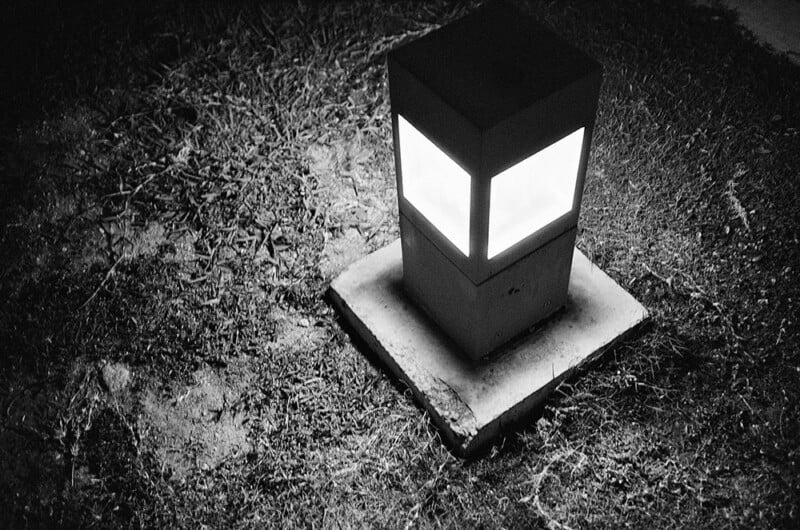 A small, square outdoor lamp emits light on a patch of grass and dirt at night. The lamp is housed in a simple, cuboid structure placed on a concrete base, casting a soft glow in the surrounding area.