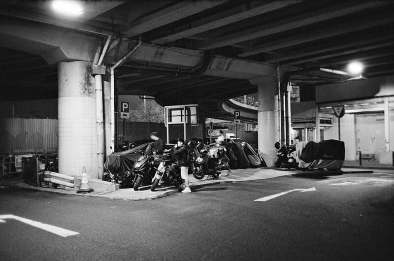 A black and white photo depicts several motorcycles parked under a highway overpass at night. Some motorcycles are covered with tarps, and a few people walk nearby. The area is dimly lit, with concrete pillars and road signs visible.