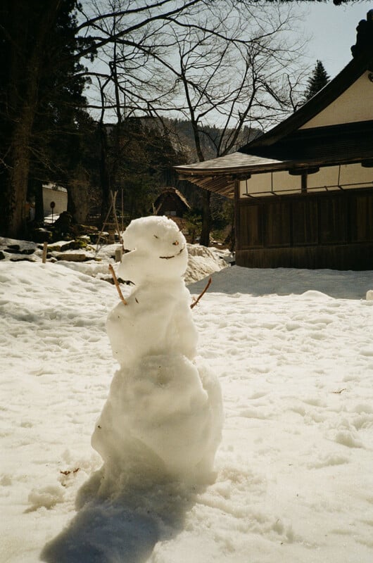 A small snowman with twig arms and a smiling face stands in a snow-covered yard. Behind it, there are leafless trees and a traditional wooden building with a sloped roof under a clear sky.