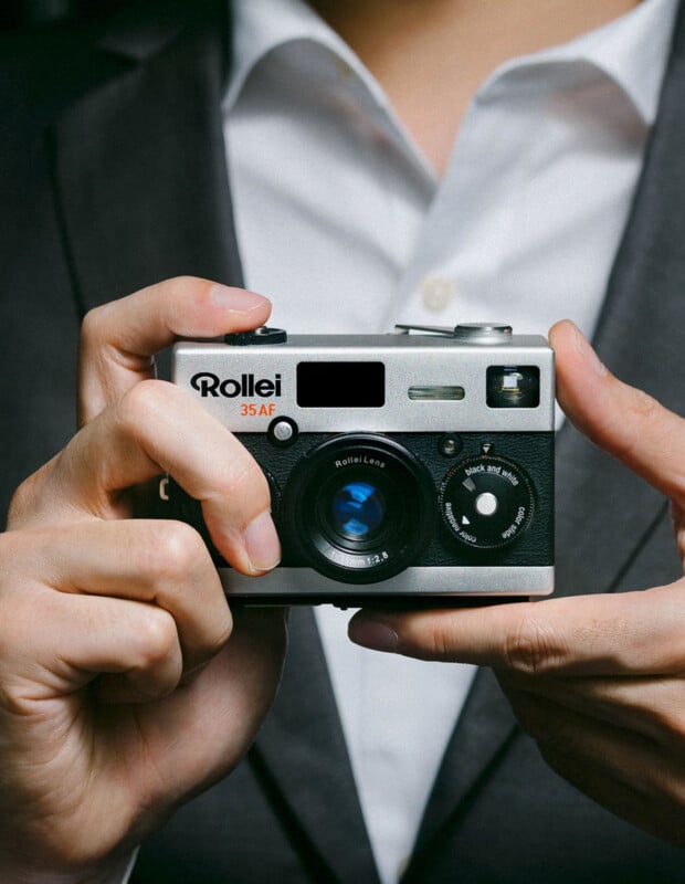 A person in a suit holds a vintage Rollei 35AF camera. The camera has a retro design with a visible lens and various control dials. The person's fingers are positioned around the camera, poised to take a photograph.