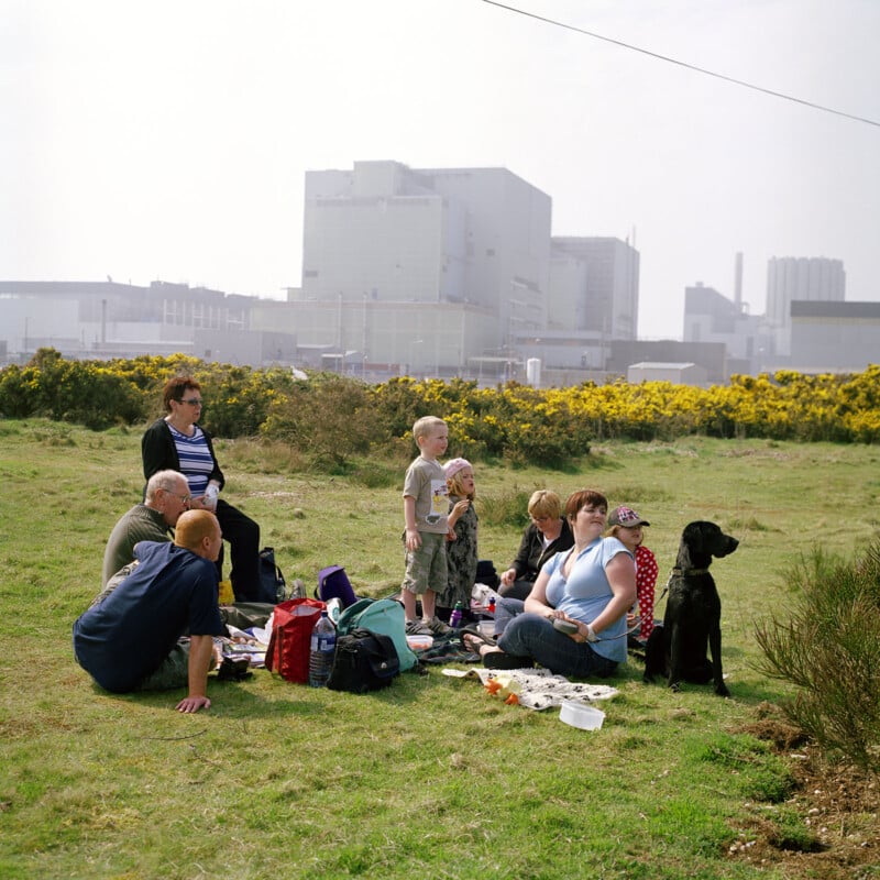 A group of people and a black dog sit on the grass, enjoying a picnic on a sunny day. Behind them is a large industrial facility with various buildings and structures. The area is surrounded by greenery and yellow flowering bushes.