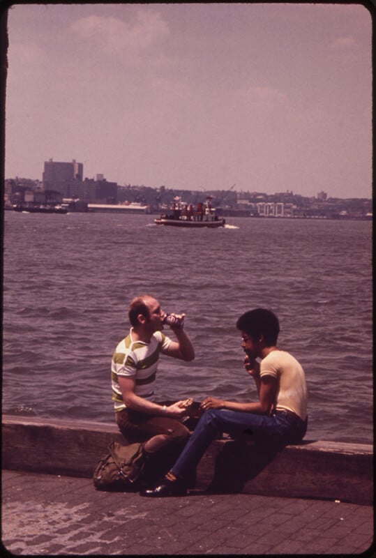 Two people sit on a bench by the waterfront, with one drinking from a can and the other eating. A river and distant city buildings are visible in the background, along with a boat on the water. The atmosphere is casual and relaxed.