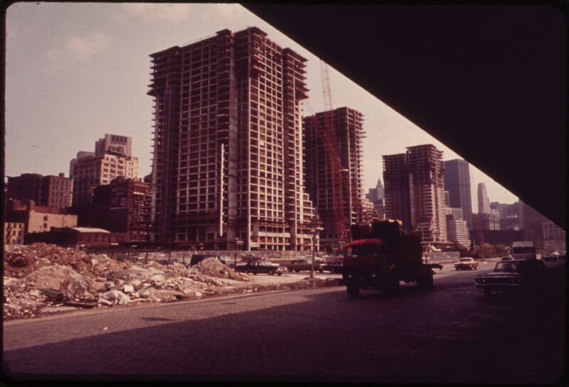An urban construction scene with tall, skeletal high-rise buildings under construction. A crane is visible along with debris and rubble in the foreground. A truck drives along the street, with other vehicles parked nearby. The city skyline is in the background.