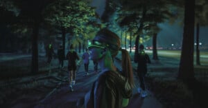 A young woman wearing futuristic neon-lit goggles walks along a dimly lit forest path at night. Several other people, also wearing neon goggles, walk or jog in the background under trees illuminated by soft, colorful lights.