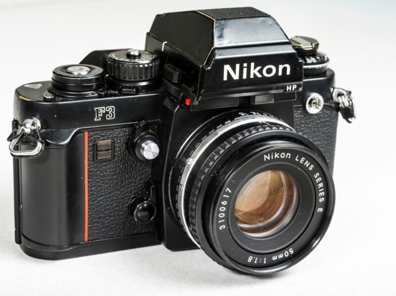 A black Nikon F3 HP film camera is shown with a 50mm Nikon lens attached. The camera has a vintage design with various dials and buttons, and features the Nikon logo prominently at the top.