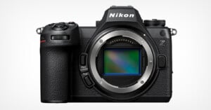 A Nikon Z series mirrorless camera body is shown against a white background. The camera, featuring a textured grip and numerous buttons, is pictured without a lens attached, revealing the sensor inside the mount. The Nikon logo is visible at the top.
