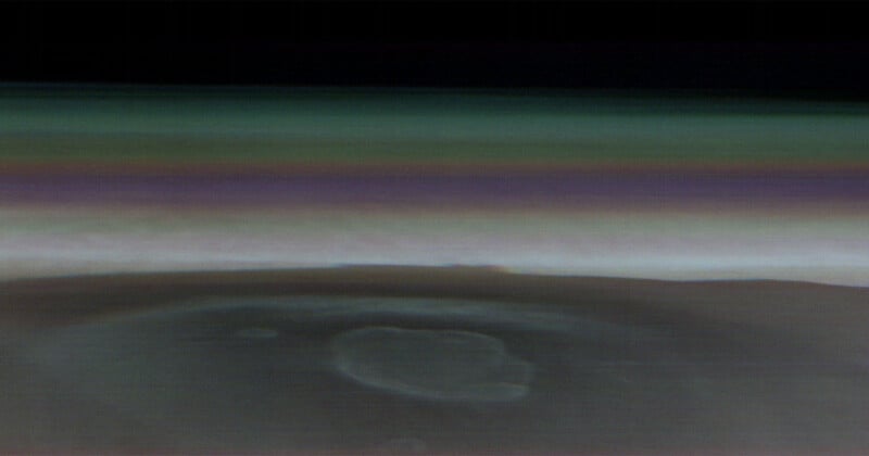A photograph depicting the upper layers of Saturn's atmosphere, taken by NASA's Cassini spacecraft. The image shows various colored bands, representing different atmospheric layers, with a large, faint circular formation visible in the lower portion.