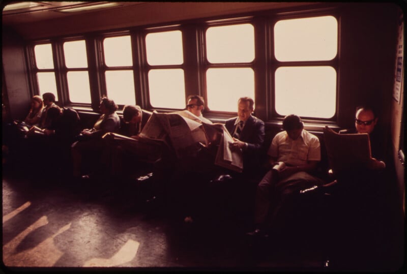 A row of passengers sitting on a train, most of them absorbed in reading newspapers. The scene is indoors with natural light coming through the large rectangular windows on the left side. The overall atmosphere is calm and quiet.