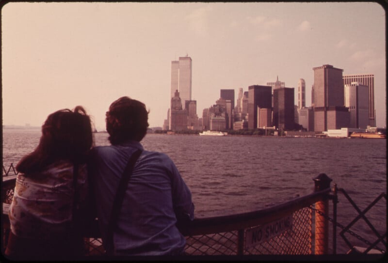 Two people are seen leaning on a railing, looking out at the New York City skyline from across the water in a vintage photo. The buildings of downtown Manhattan, including the Twin Towers, are prominent against a slightly hazy sky.