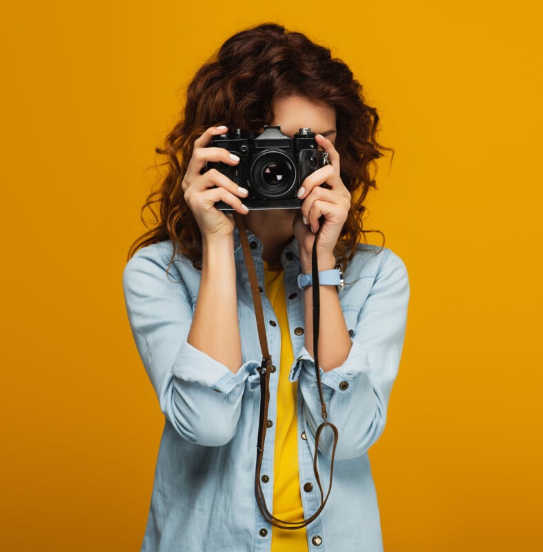 A person with curly hair wearing a light blue denim shirt is holding a black camera up to their face against a bright yellow background. The camera's strap is hanging down from their hands.
