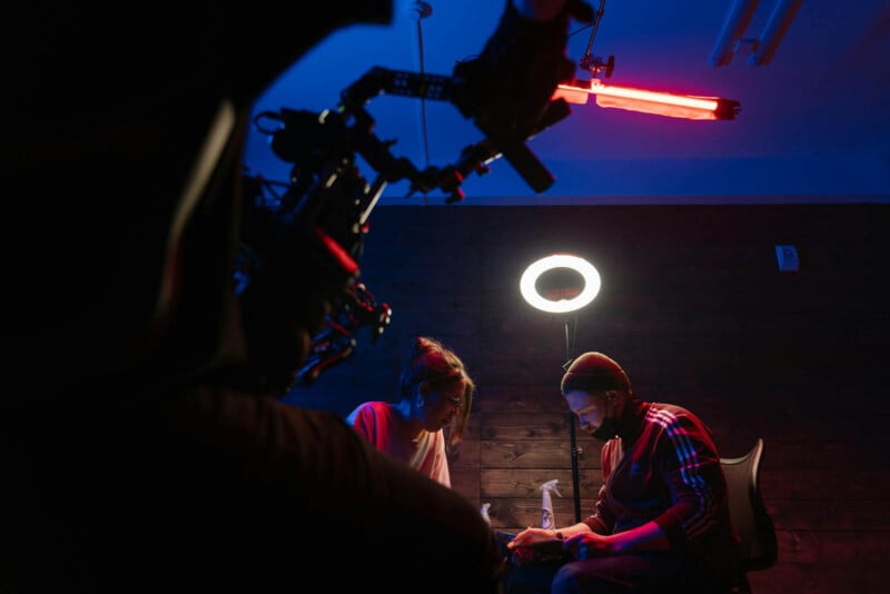 Two people seated at a table under a circular light, engaged in a task. The room is dimly lit with blue and red lighting. A camera rig is positioned in the foreground, framing the scene.