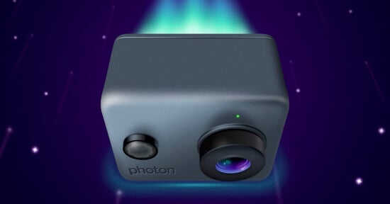 A compact, dark gray camera hovers against a space-themed background with streaks resembling stars. The camera has the word "photon" inscribed on the front, with a prominent lens and a small green light above it. A soft glow emanates from behind.