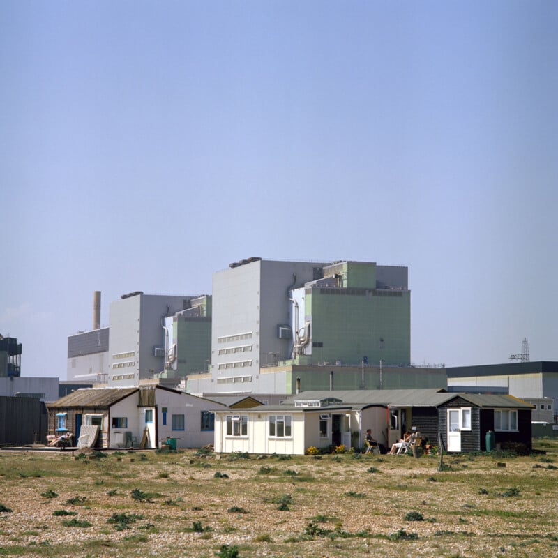 A large industrial facility with multiple interconnected large, grey buildings is in the background. In the foreground, there are several small, simple houses or cottages on sparsely vegetated land. The sky above is clear and blue.