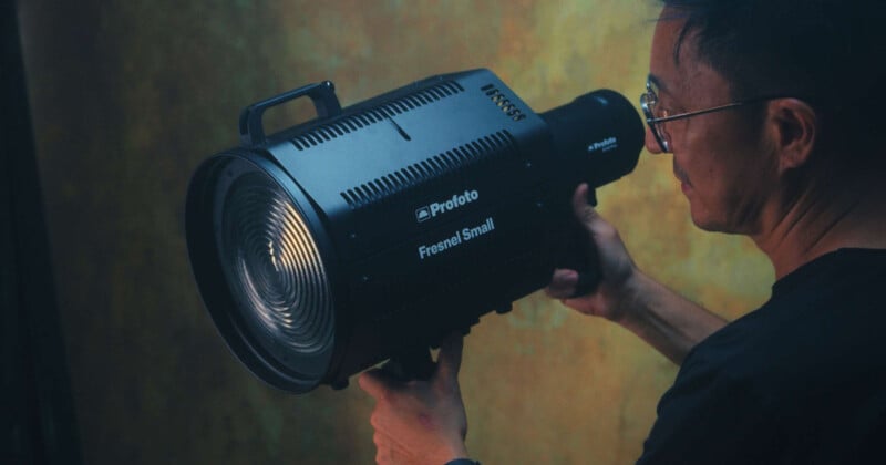 A person wearing glasses holds a Profoto Fresnel Small lighting unit, preparing to adjust or position it. The background is dimly lit with a mottled pattern, creating a dramatic and focused atmosphere.