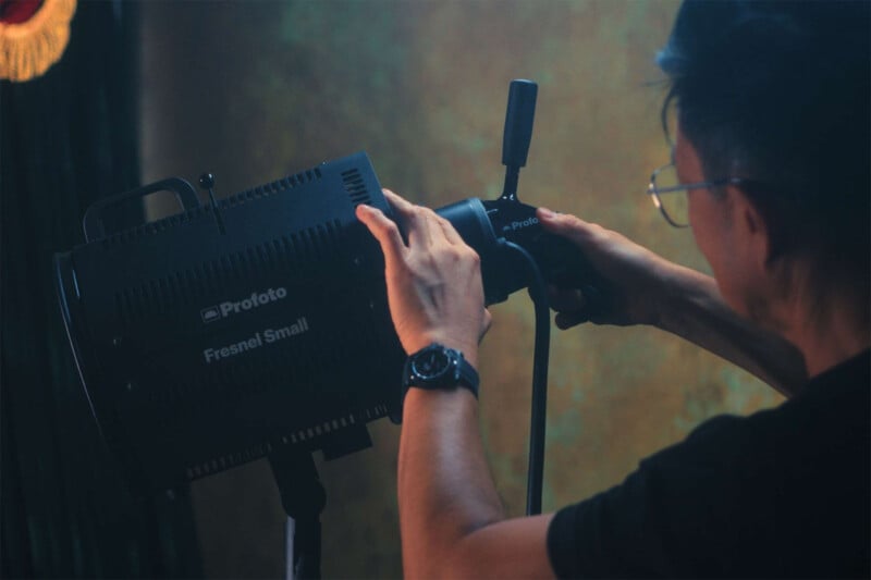 A person wearing glasses adjusts a Profoto Fresnel Small studio light. The individual is using both hands to fine-tune the light's position. The background is softly lit, creating a professional studio atmosphere.