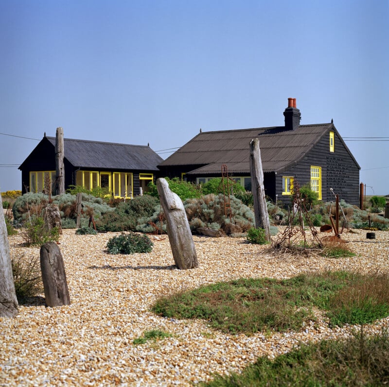 A charming black wooden house with bold yellow window frames stands in a gravel garden adorned with sparse greenery and tall, weathered wooden posts. The scene is set against a clear blue sky, giving a rustic, coastal vibe.