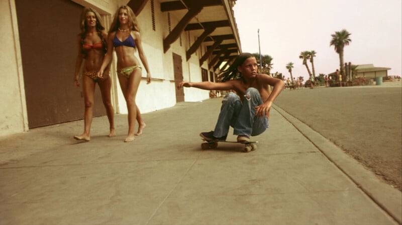 A person is crouched on a skateboard, riding on a wide sidewalk along a beach. Two women in bikinis and beachwear walk nearby. The environment features a sandy area, palm trees, and an old building with a wooden roof structure. The sky appears clear with a warm hue.