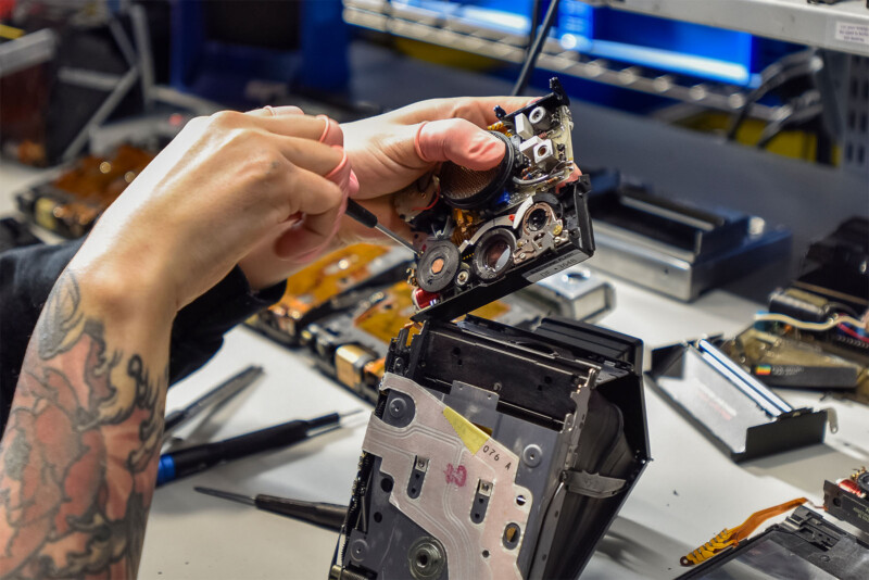 A person with tattooed arms is disassembling a camera, with various small tools and electronic components scattered on the workbench. The background includes shelves with more electronic equipment and a computer screen.