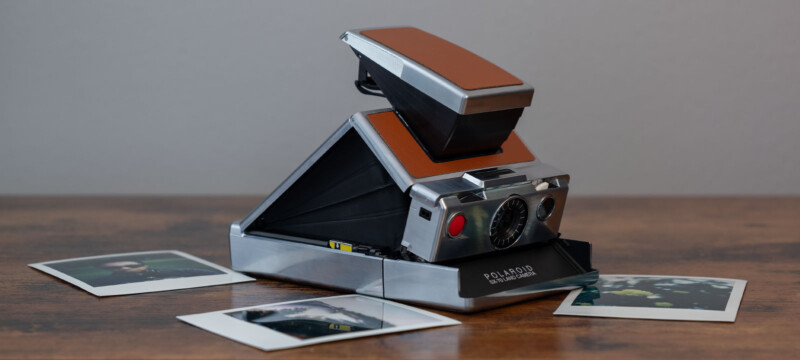 A vintage Polaroid SX-70 Land Camera with a brown leather exterior sits on a wooden surface. The camera is partially unfolded. Two instant photographs are scattered in the foreground, partially visible. The background is a plain gray wall.