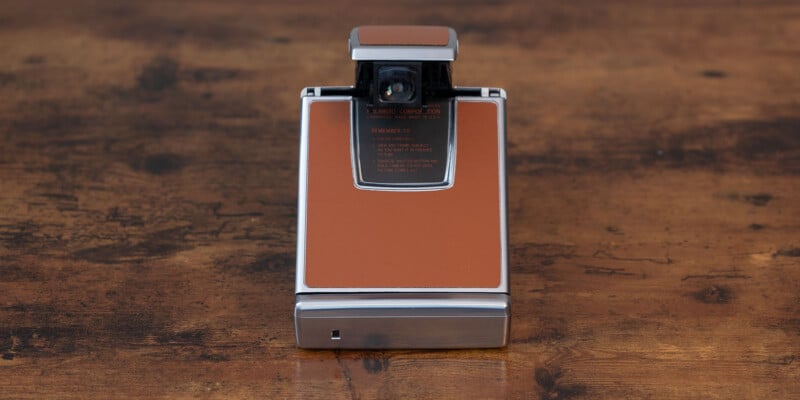 A vintage Polaroid SX-70 Land Camera with a brown leather exterior and silver metal frame is placed on a wooden surface. The camera is in a closed position, showing its rectangular shape and sleek design.