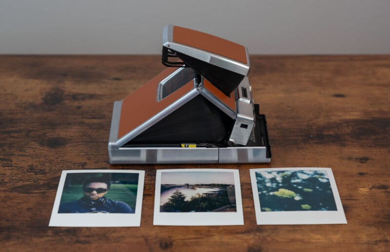 A vintage instant camera with a brown exterior sits on a wooden surface. In front of it are three polaroid photos: one of a person wearing sunglasses, one of a scenic view with a body of water, and one of yellow flowers in the foreground.