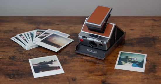 A vintage Polaroid Land Camera sits on a wooden surface. To the left, there is a spread of Polaroid photos, and two additional photos lie in the foreground. The images show landscapes and a dog. The scene is cozy and nostalgic.