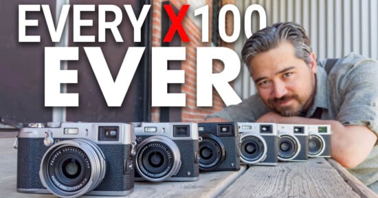 A person with grey hair and a beard is leaning on a wooden table, smiling at the camera. In front of them, there are six different Fujifilm X100 series cameras lined up. The text above reads "EVERY X100 EVER." The background has an industrial look with brick and metal elements.
