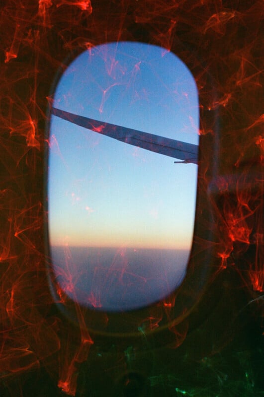 View of an airplane wing through a window at high altitude with a gradient sky in the background, transitioning from blue to orange near the horizon. The scene is overlaid with vibrant red and green light streaks swirling around the edges.