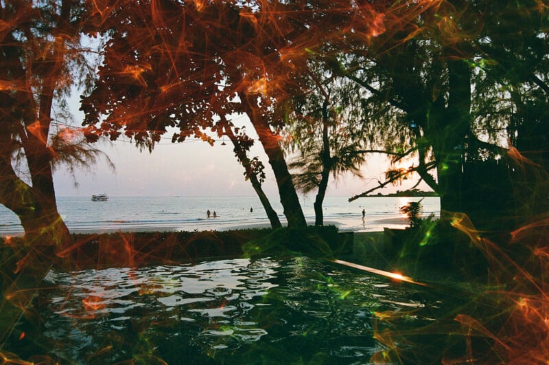 An enchanting beach scene at sunset, viewed through trees lining the shore. Silhouettes of people are seen in the distance near the water. The foreground features reflections in what appears to be a pool, with vibrant light streaks enhancing the image's atmosphere.