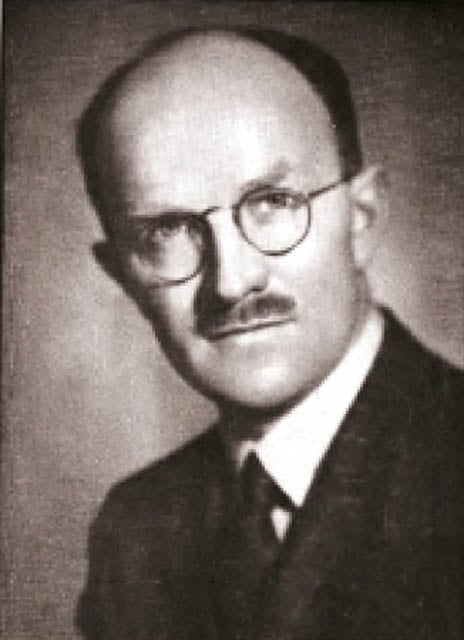 Black and white portrait of a man with a bald head and neatly trimmed mustache, wearing a dark suit, white shirt, and tie. He wears round glasses and faces the camera with a serious expression. The background is plain and blurred.