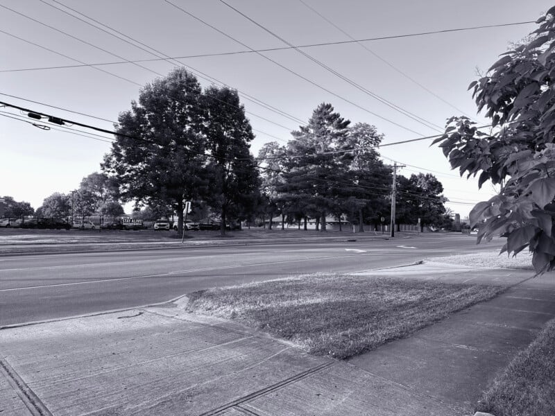 A monochrome image shows a tranquil suburban street with neatly trimmed grass on either side. Trees and power lines line the street, and a few parked vehicles are visible in the distance. The sky above is clear, indicating fair weather.