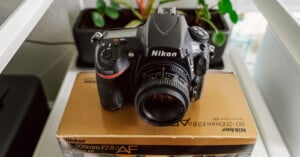 A Nikon camera with an attached lens is placed on top of a Nikon Nikkor 80-200mm f/2.8D AF lens box. The background includes a blurred view of plants and a shelf, adding a touch of greenery to the setup.