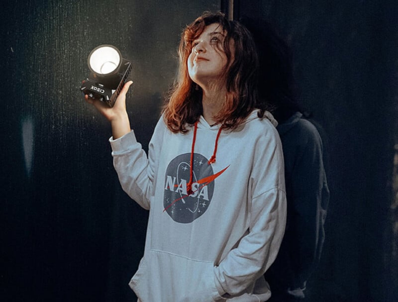 A person with shoulder-length hair is standing in a dimly lit room, wearing a white NASA hoodie. They are holding up a lit spotlight with a contented expression, casting a glow on their face and part of their surroundings.