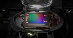 A close-up view of a camera sensor inside a device, with a lens positioned above it. The multicolored surface of the sensor is clearly visible, surrounded by intricate electronic components and a metal frame. The background is dark, highlighting the sensor.