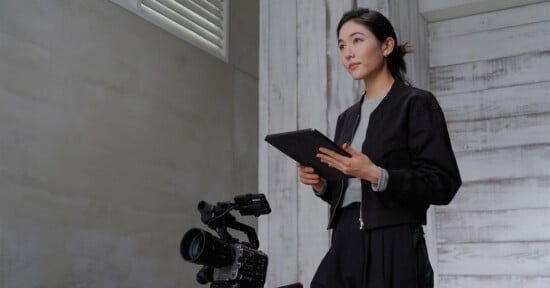 A woman holding a tablet stands next to a professional video camera in a modern, industrial-style room. She is wearing a dark jacket and appears focused, possibly preparing for a video shoot or production task.