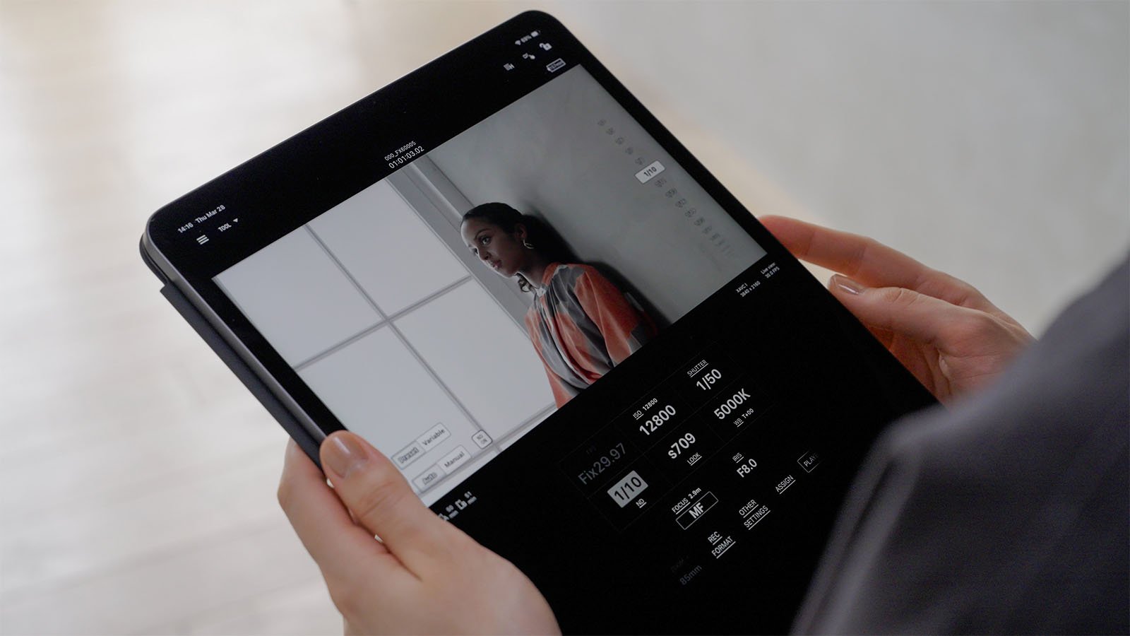 A person holds a tablet displaying a photo editing interface. The screen shows an image of a person standing by a window, wearing a striped shirt. Editing options and settings, such as ISO, white balance, and shutter speed, are visible on the tablet screen.
